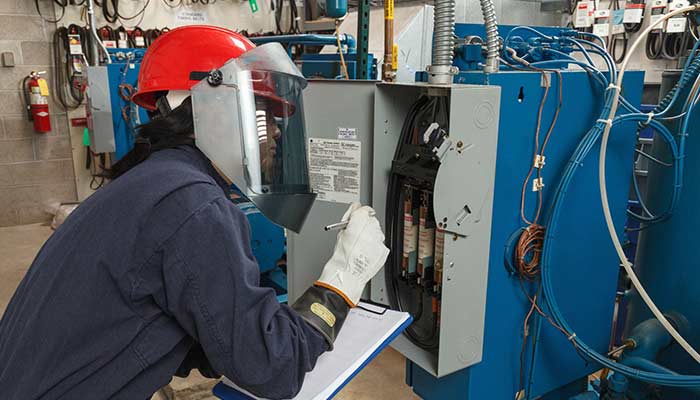 A worker wearing heavy safety gear inspects a panel that contains arc flash compressors.