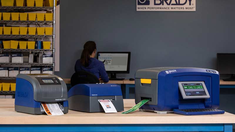 Three different Brady label printers showcasing safety labels in an office environment.