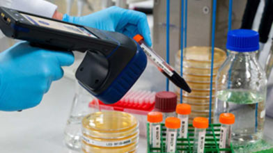 Scientist in a lab coat extracting a vial from a tube rotator and scanning it with an RFID reader, surrounded by laboratory equipment.