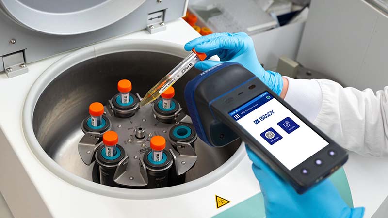 Scientist in a lab coat extracting a vial from a tube rotator and scanning it with an RFID reader, surrounded by laboratory equipment.