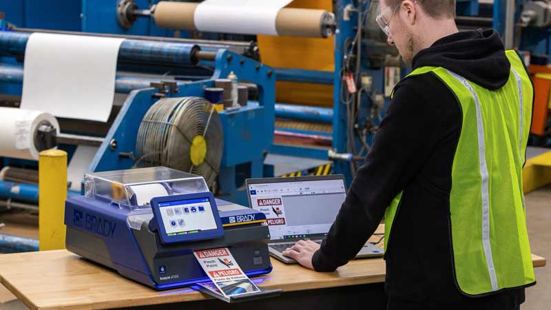 Contractor printing a "danger, pinch point" label with a Brady J7300 inkjet benchtop printer in a warehouse.